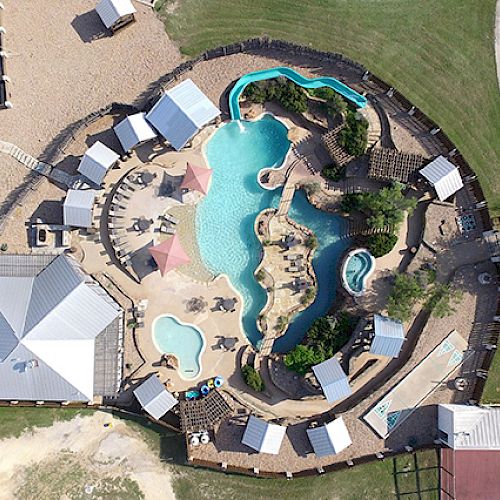 Aerial view of a swimming pool complex with umbrellas, lounge chairs, cabanas, and landscaped greenery, surrounded by a parking lot.