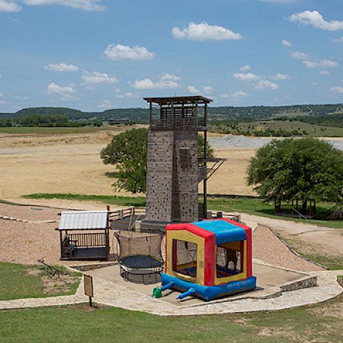 An outdoor recreational area featuring a rock-climbing tower, a bounce house, and some shaded structures with a scenic backdrop, visible.