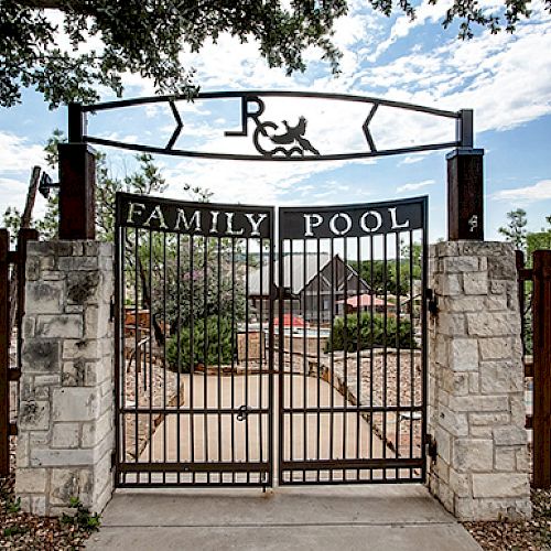 This image shows a gated entrance to a family pool area. The gate is flanked by stone pillars and a wooden fence, with trees and a clear sky behind it.