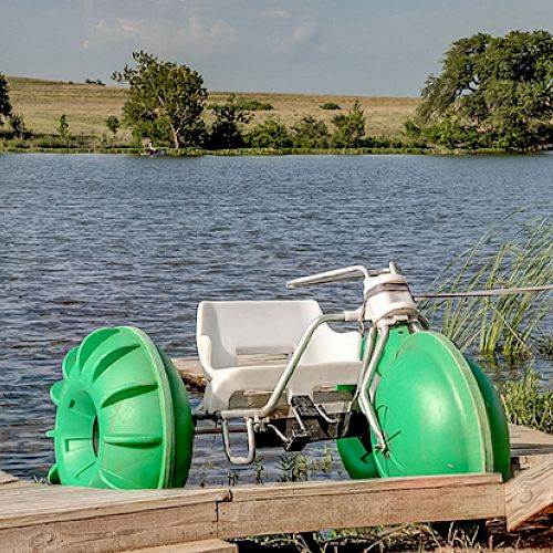 A green pedal boat with large wheels is parked on a wooden dock by a lake, surrounded by grassy landscape and trees.