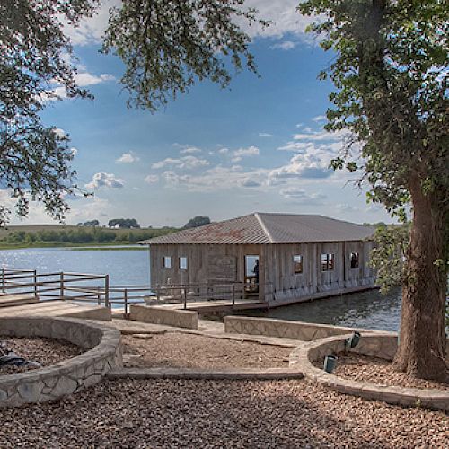 The image shows a small wooden building on a lake, surrounded by trees, with a dock and greenery under a partly cloudy sky.