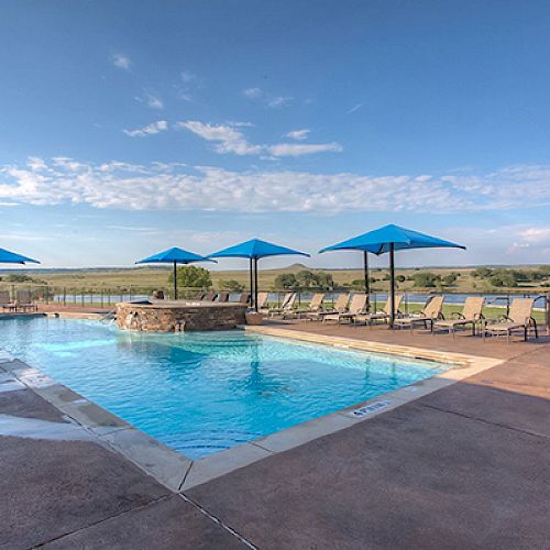 The image shows an outdoor swimming pool with blue umbrellas and sun loungers around it, set against a scenic background of rolling landscape.