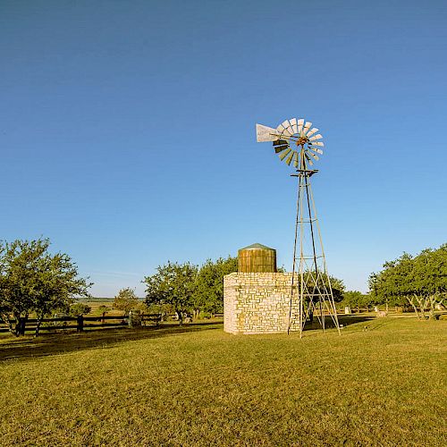 The image shows a windmill next to a stone structure in an open field with scattered trees and a clear blue sky in the background.