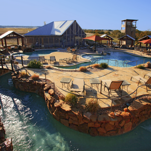 The image shows a serene outdoor pool area with lounge chairs, a lazy river, and a rustic building under clear blue skies.