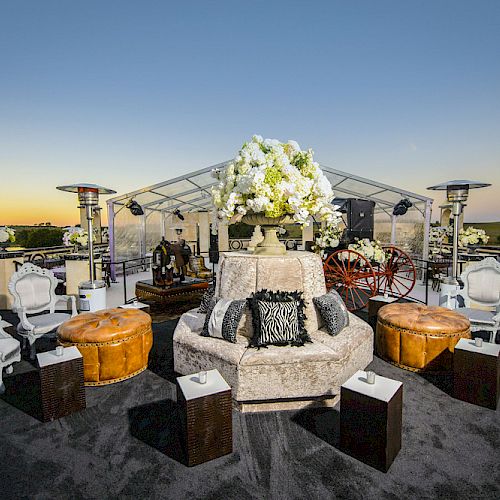 An elegant outdoor seating area featuring white chairs, plush ottomans, a circular sofa with floral centerpiece under a clear tent at sunset.