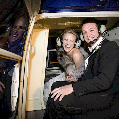 Two people dressed in wedding attire, smiling, and sitting inside a helicopter with headsets on, possibly during a celebratory moment.