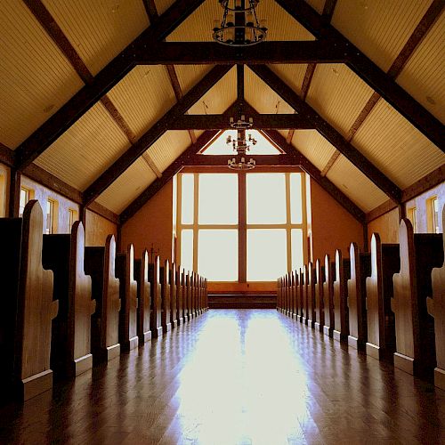 An empty church interior with wooden pews on both sides, a high ceiling, and large windows at the front allowing natural light in.