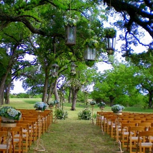 The image shows an outdoor wedding setup with rows of wooden chairs, floral arrangements, and lanterns hanging from trees, in a lush, green environment.