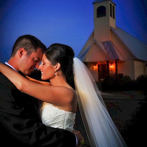 A newlywed couple lovingly embraces outside a church at twilight, both in formal wedding attire, with soft lighting highlighting their faces.