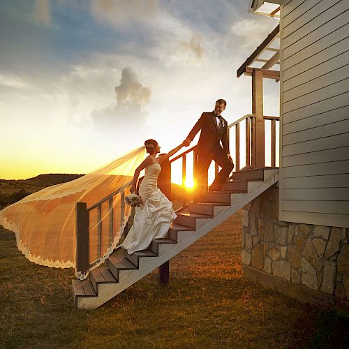 A bride and groom hold hands as they descend outdoor steps near a house, with the sun setting in the background and her veil flowing behind.