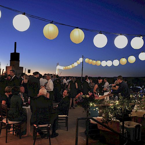 An outdoor evening event with people socializing under string lights and paper lanterns, against a twilight sky.