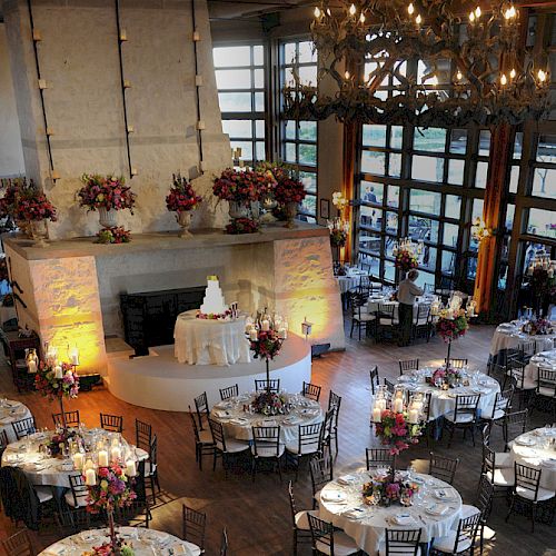 An elegant indoor reception area with round tables set for an event, featuring a central cake display and a large fireplace decoration.