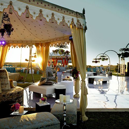 Outdoor event setup with elegant tents, draped curtains, comfortable seating, decorative lights, and a reflective flooring surface during sunset.