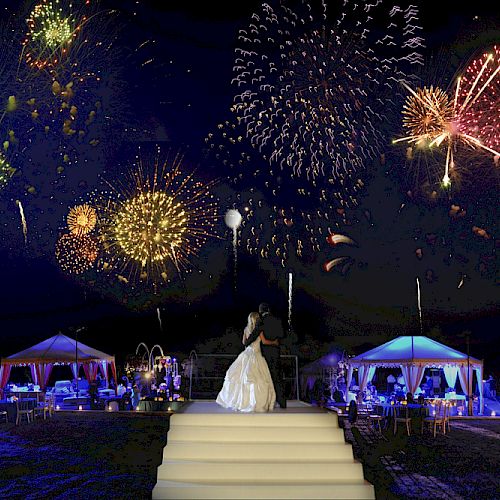 A couple stands on steps, facing a vibrant display of fireworks under a night sky, amidst illuminated tents and tables in a celebratory setting.