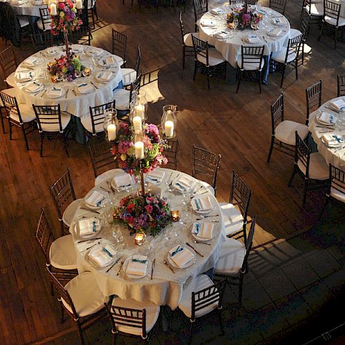 An elegant event setup featuring round tables with white tablecloths, floral centerpieces, and place settings in a warmly lit room with wooden floors.