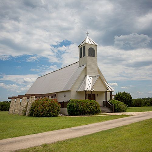 The image depicts a small white church with a bell tower, surrounded by green grass, bushes, and trees, under a partly cloudy sky.