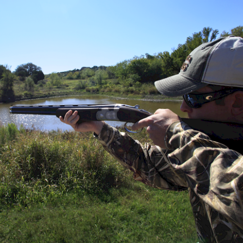 A person in camouflage aims a shotgun near a lake with trees and greenery in the background, wearing a cap and sunglasses.