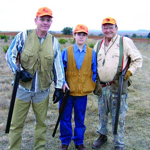 Three people wearing hunting gear and orange hats stand outdoors holding rifles. They are in a grassy, open area with a trail in the background.