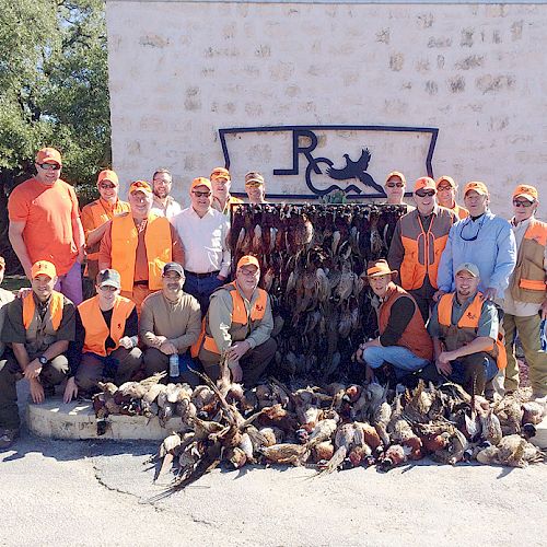 A group of people dressed in hunting gear poses in front of a wall with birds they hunted, with a sign featuring a dog above them.