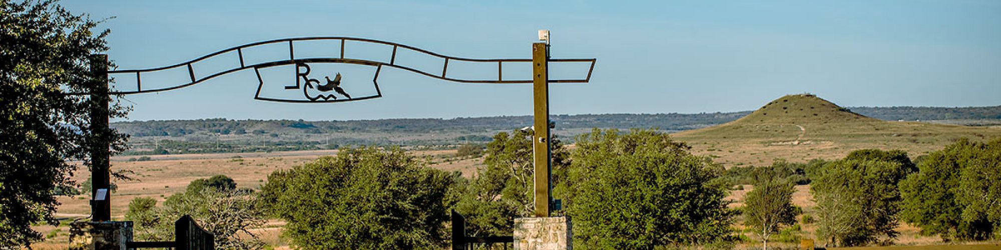 A rural landscape features a ranch entrance with a metal archway, trees, a distant hill, and vast open fields under a clear blue sky.