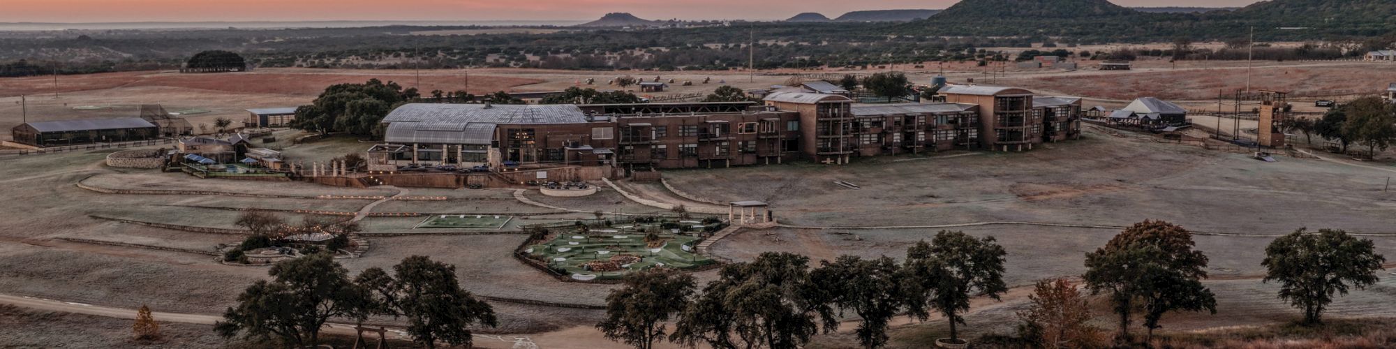 Aerial view of an industrial/campus complex with brick buildings and green roofs beside a river at sunset.