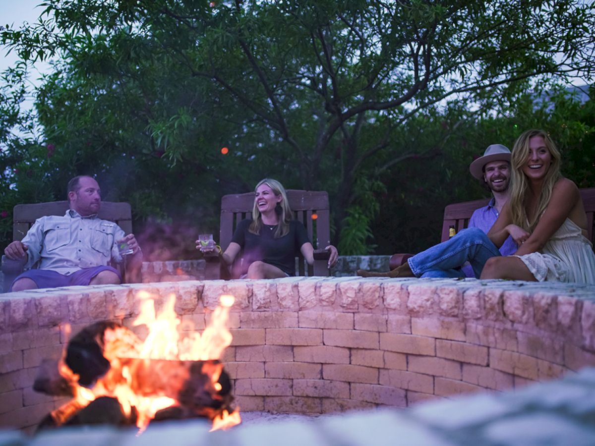 Four people are sitting around a brick fire pit outdoors, enjoying a fire. Two are seated on the left, and a couple is on the right, smiling.