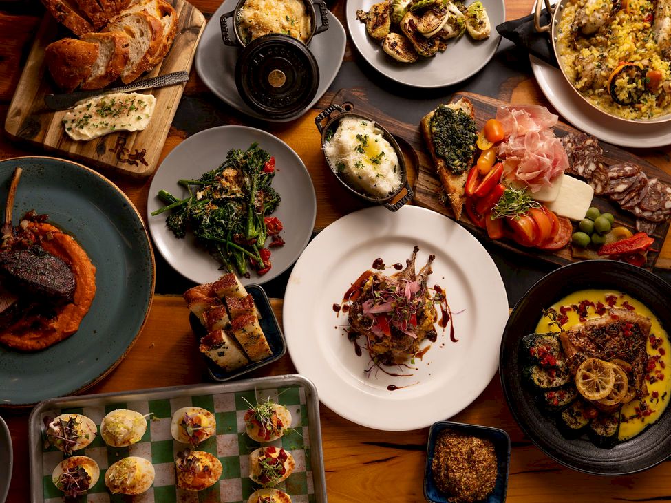 A tabletop spread of gourmet foods: bread, seafood, roasted meats, salads, polenta, and pizza-like dishes.