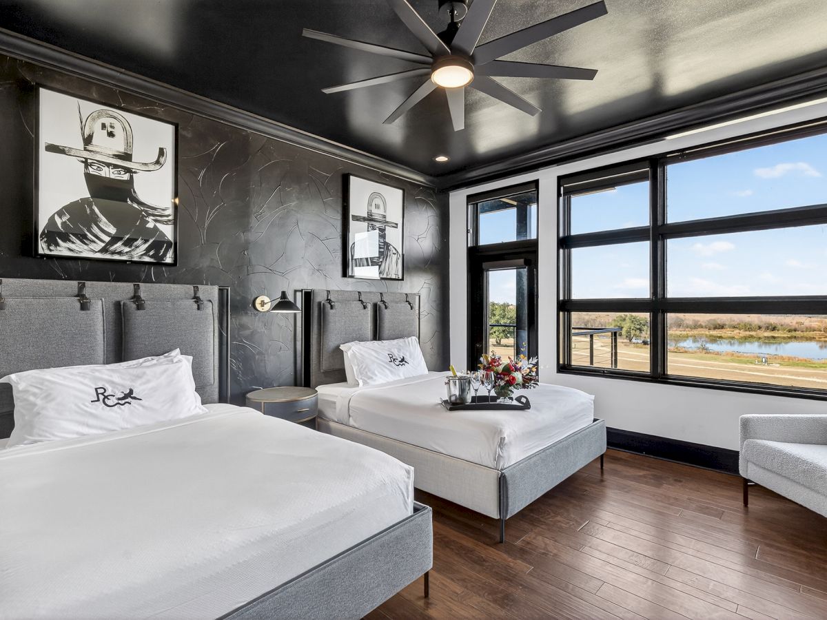Modern bedroom with two gray twin beds, a ceiling fan, cowboy wall art, and large windows overlooking a river and field.