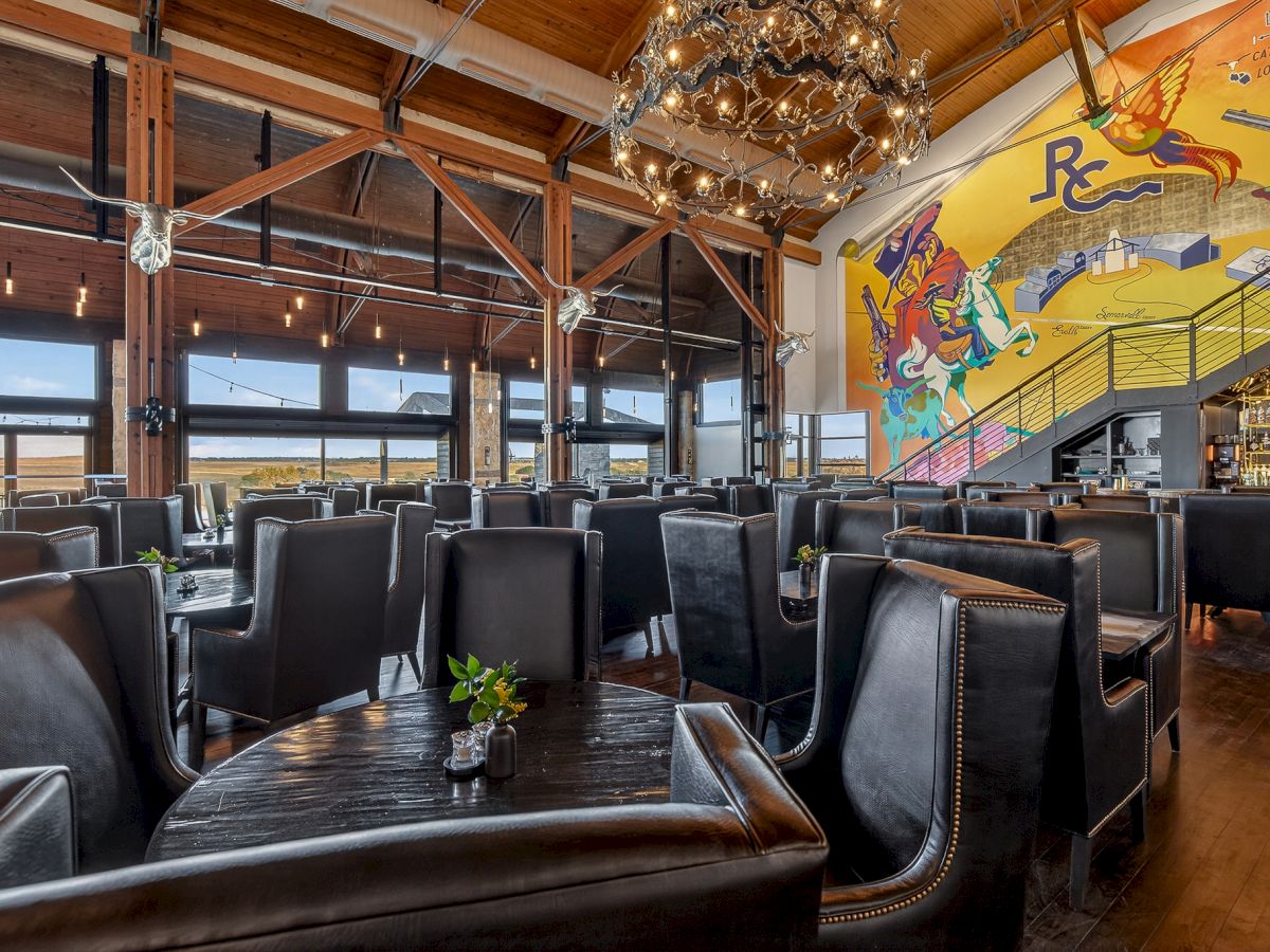 A rustic bar lounge with black leather seating, warm chandelier lights, cowboy-themed mural, and a drinks counter in a wood-ceiling room.