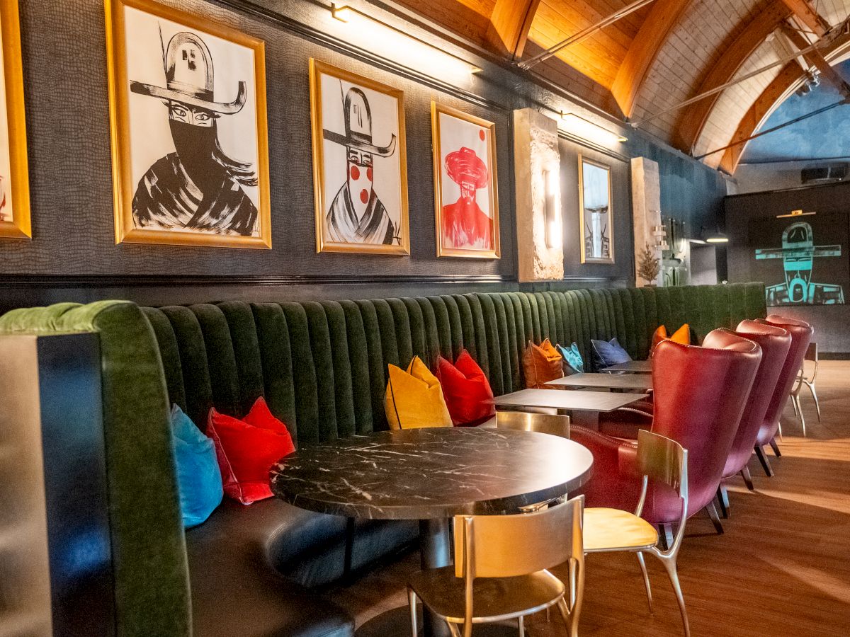 A cozy modern lounge or caf&eacute; interior with tufted green seating, colorful pillows, and framed art on dark walls.