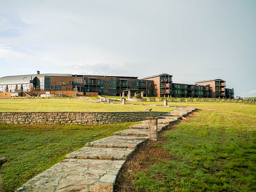 The image shows a modern building complex with a stone pathway leading up to it, set against a grassy landscape under a cloudy sky.