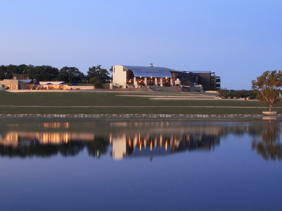 A large building with a blue roof sits beside a calm pond or lake, lights glowing over a grassy lawn with mirrored reflections.