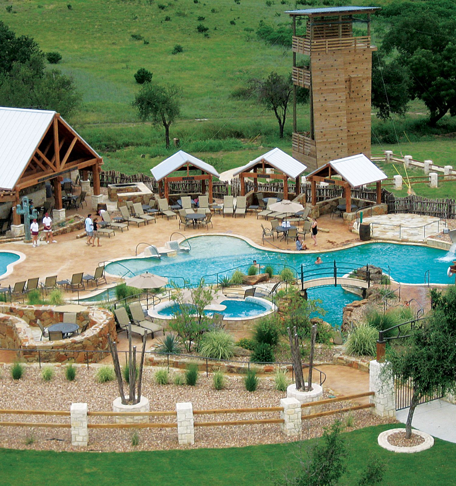 The image shows a resort area featuring pools, a hot tub, seating areas, a wooden structure, and surrounding greenery, perfect for relaxation.
