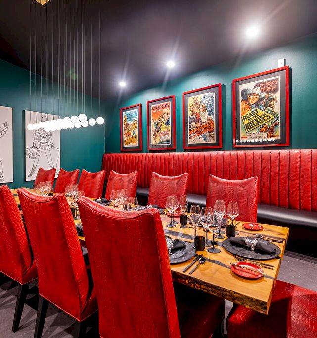 A stylish private dining room with red upholstered booth seating, wooden table settings, and framed movie posters on the wall.