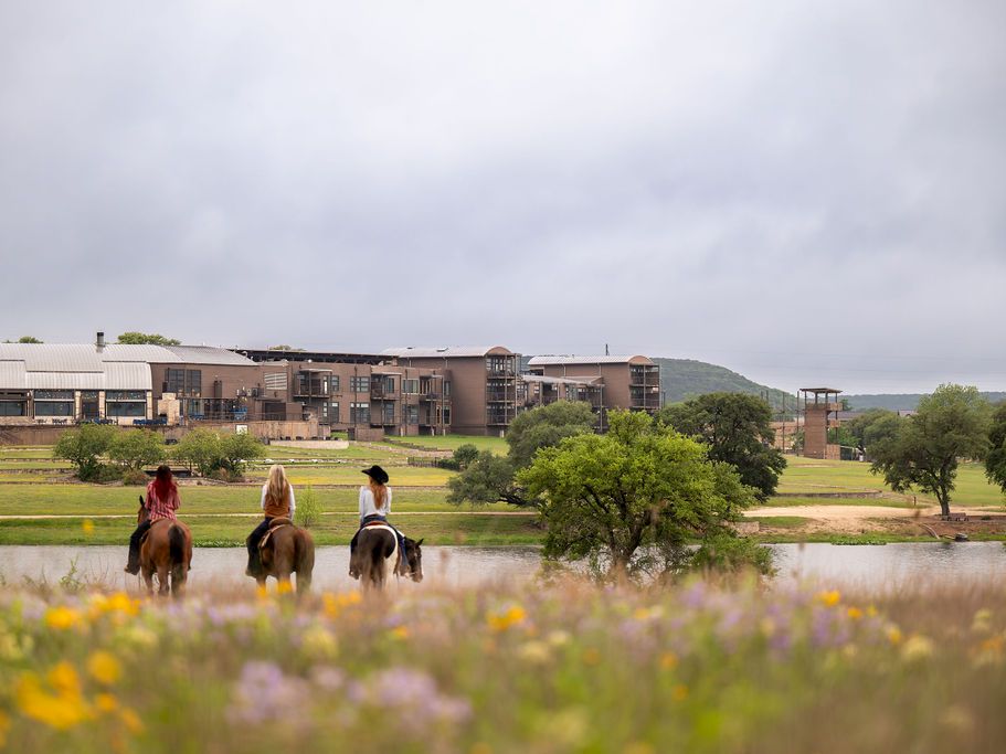 Three people ride horses along a pond in front of a landscaped campus of modern buildings under a cloudy sky.