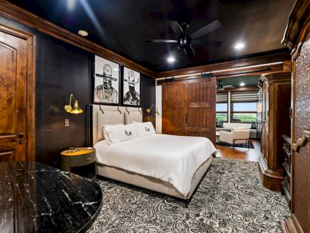 A cozy, upscale bedroom with a large white bed, dark wood walls, patterned carpet, ceiling fan, and warm bedside lighting.