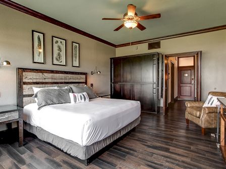 A well-decorated bedroom with a large bed, artwork above, wooden flooring, a ceiling fan, and a chair in the corner.