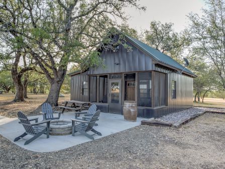 The image shows a small rustic cabin with a green roof and a small patio with several chairs around a firepit surrounded by trees.