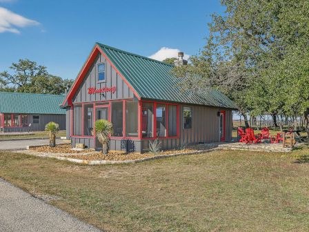 A small wooden building with a green metal roof and red trim sits beside a road, surrounded by trees and a sunny, open yard.