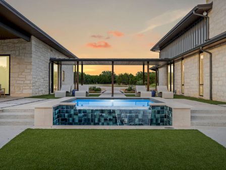 A modern stone courtyard at sunset with an outdoor hot tub/pool, lounge chairs, and a manicured green lawn, overlooking trees.