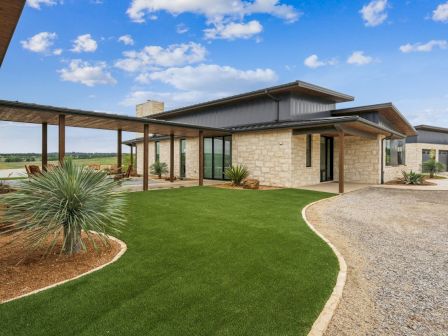 Modern house with stone walls, large windows, and a metal roof, surrounded by a landscaped yard with a clear sky overhead.