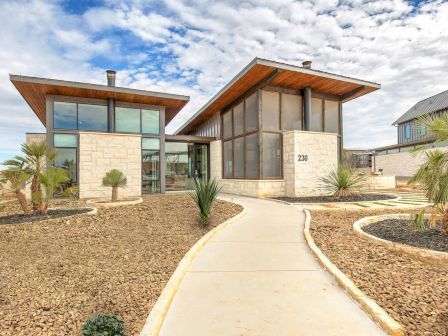 A modern building with large windows, stone walls, and a sloped roof, surrounded by a desert landscape and a concrete path leading to the entrance.