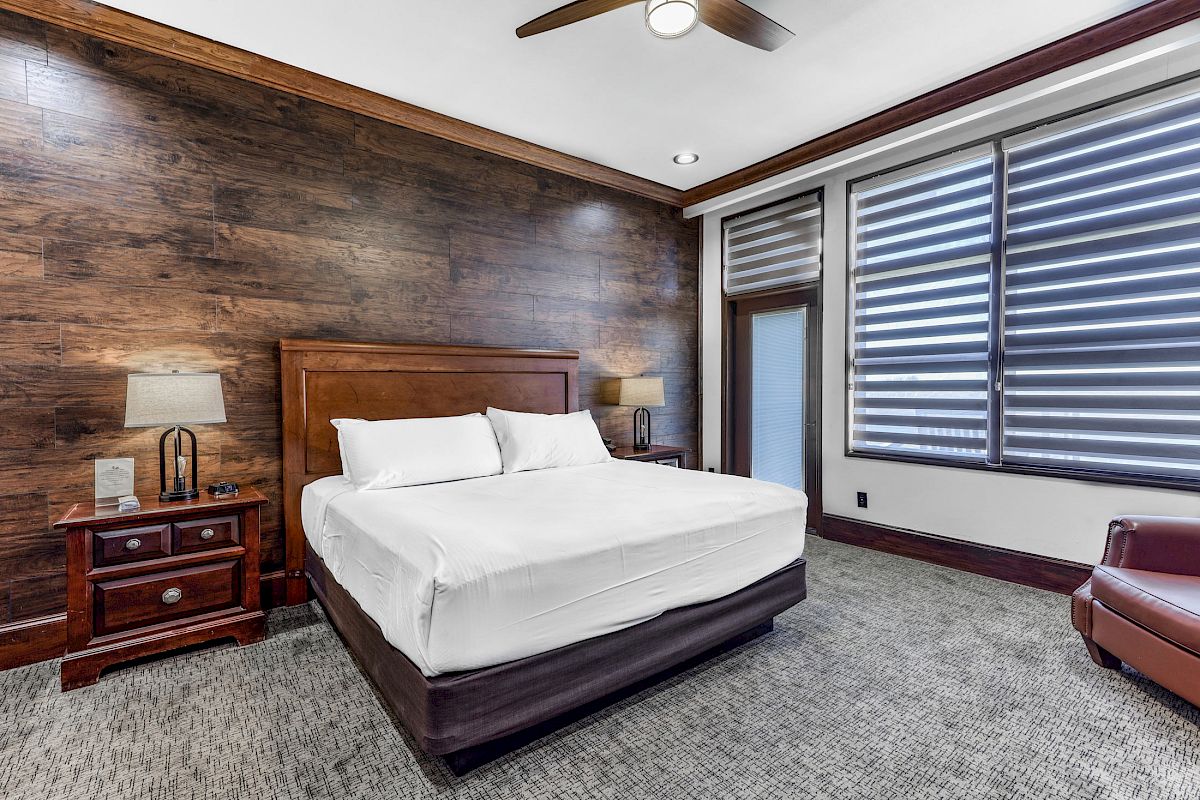 A modern bedroom with a wooden accent wall, a large bed, two nightstands with lamps, large windows with blinds, and a ceiling fan.