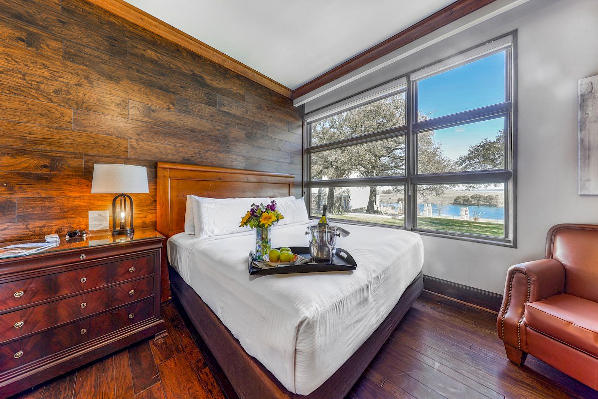 A cozy hotel room with a neatly made bed, a tray with flowers and drinks, a large window with a scenic view, and a leather armchair.