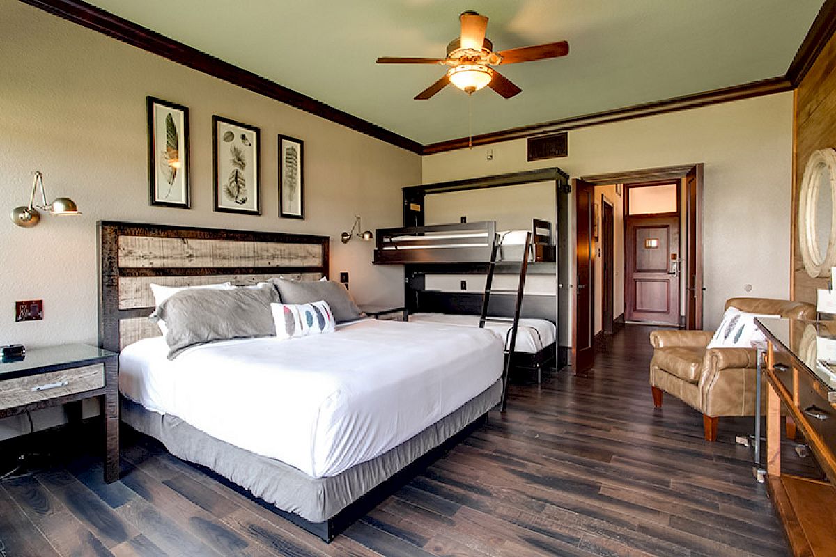 A cozy hotel room with a large bed, bunk beds, a ceiling fan, and modern decor, complemented by a wooden floor and soft lighting.