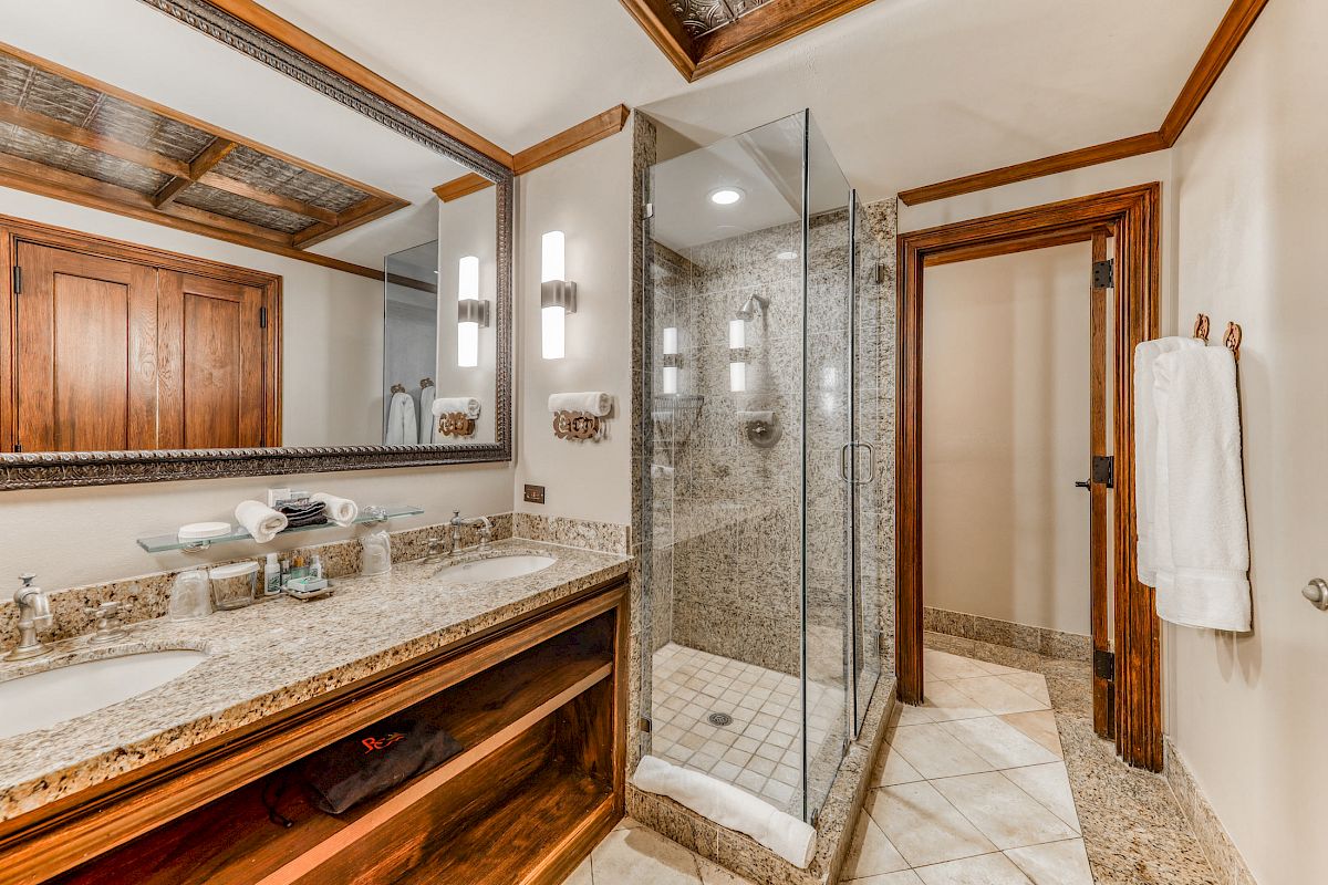 The image shows a bathroom with a granite countertop, dual sinks, a large mirror, a glass-enclosed shower, towels, and wooden doors with intricate detailing.