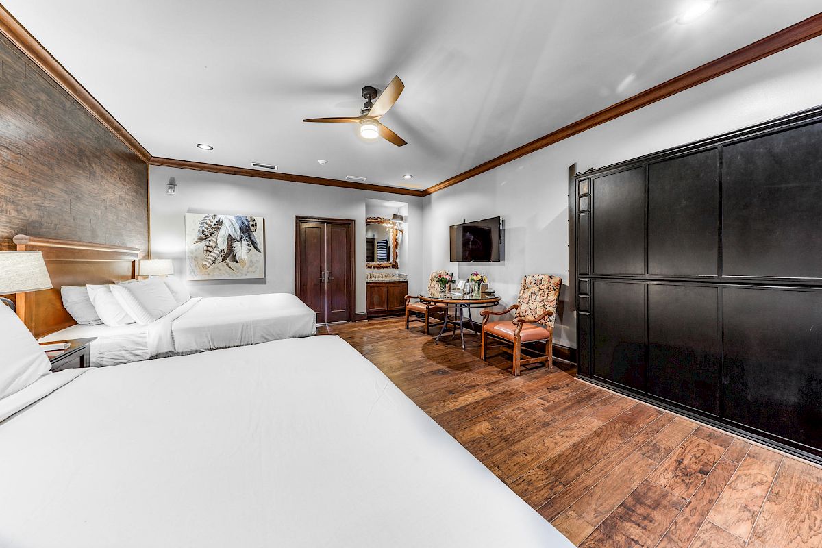 The image shows a hotel room with two beds, a ceiling fan, a wall-mounted TV, a painting, a wooden floor, and a seating area with chairs and a table.