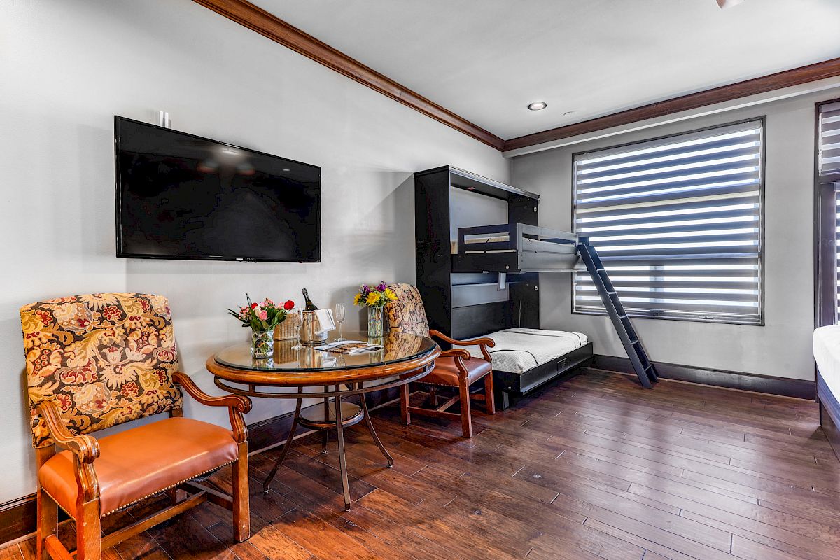 A room with a flat-screen TV, two chairs, a small round table with flowers and wine, and bunk beds near a window with striped blinds.