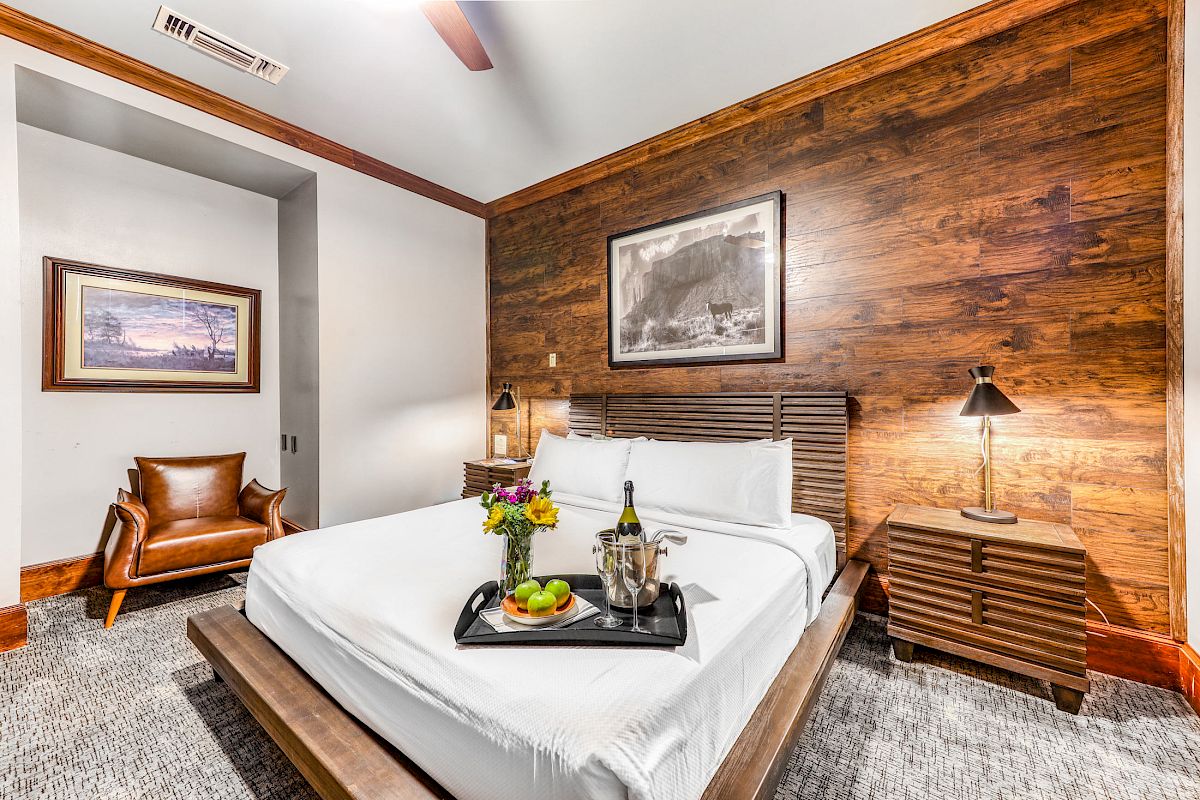 A cozy bedroom with a comfortable bed, wooden accent wall, bedside lamps, framed art, a leather chair, and a tray with flowers, fruit, and wine.