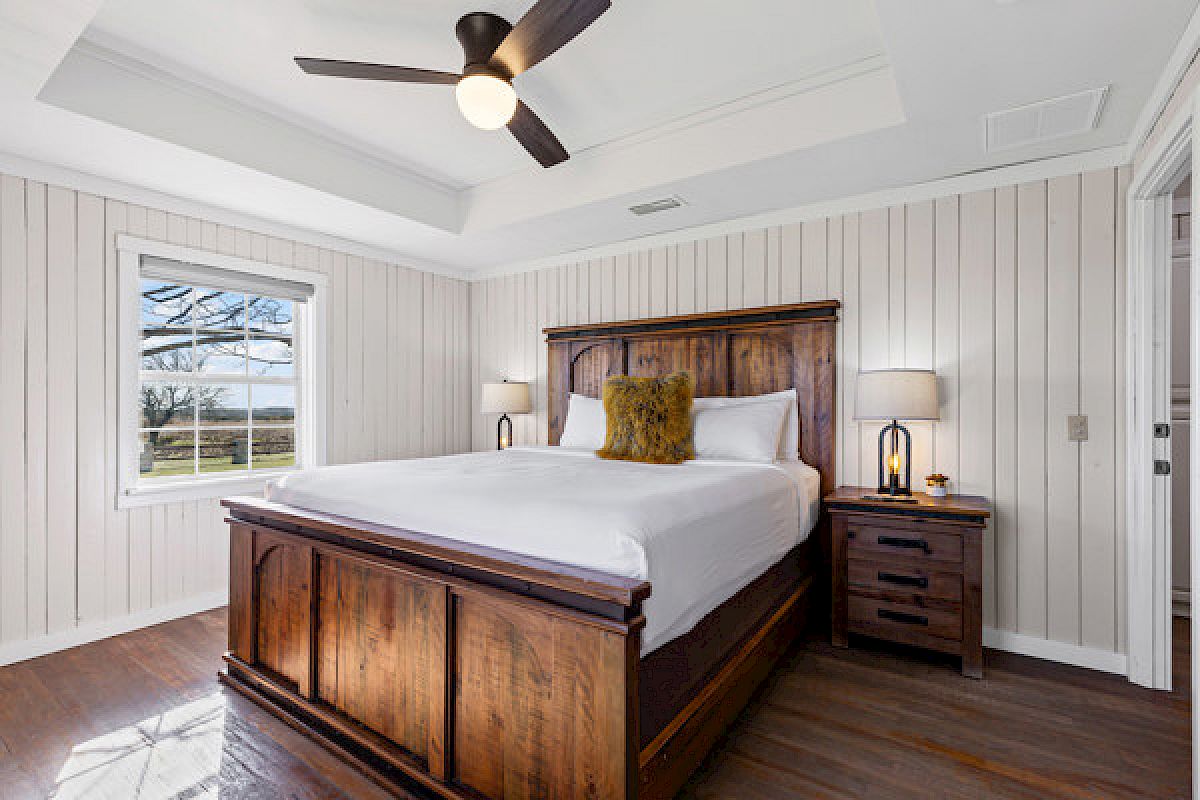 A cozy bedroom with a wooden bed, white bedding, nightstands, lamps, a window with a view, and a ceiling fan. Wood paneling adds warmth.